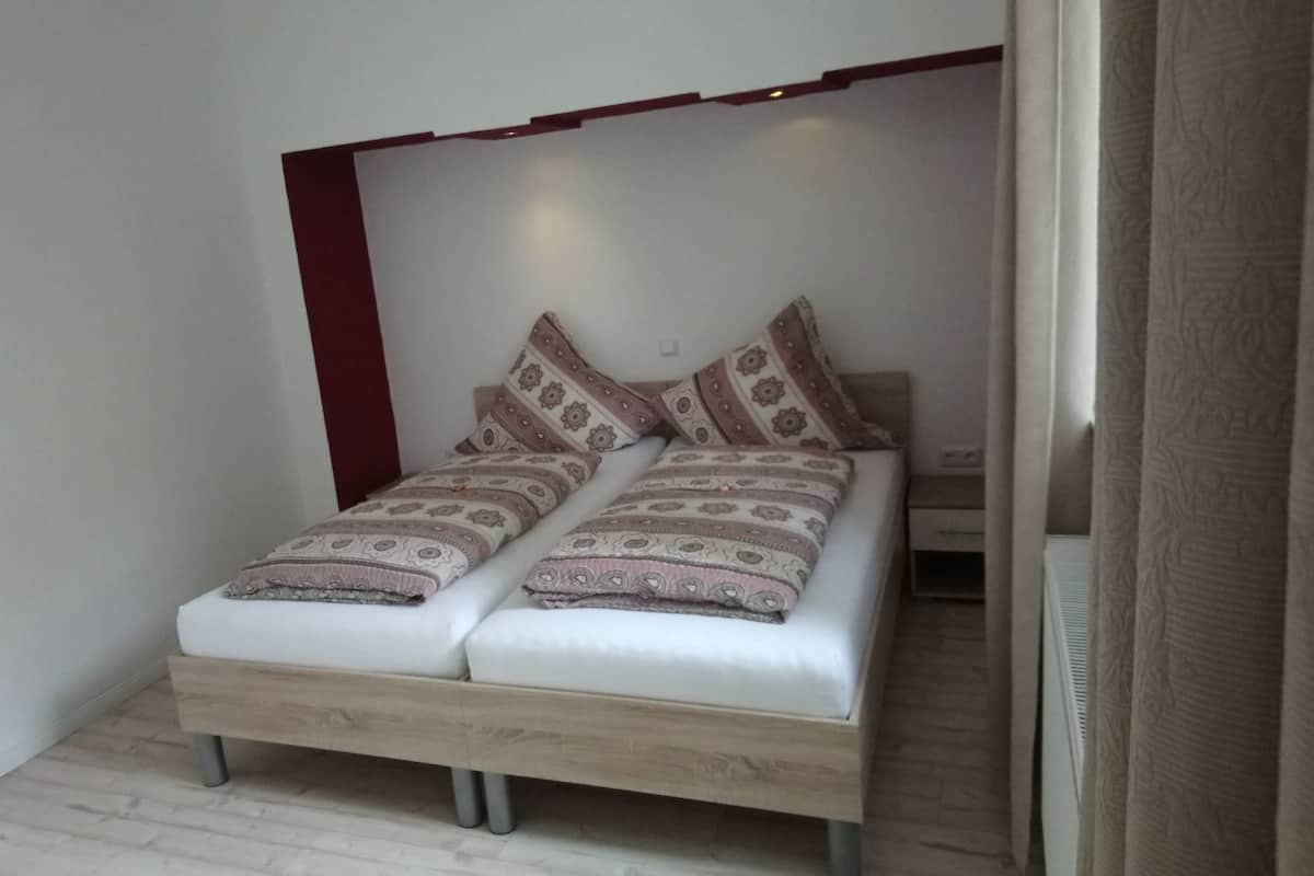 A small, single-bed room with a wooden bed frame, patterned bedding, and a red accent wall.