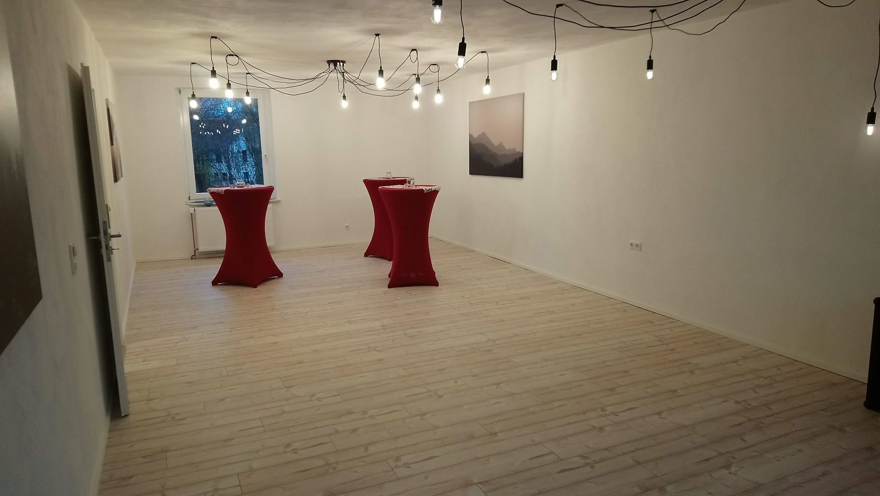 An empty room with wooden flooring, two red tables, a window with a view, and a chandelier with multiple bulbs.