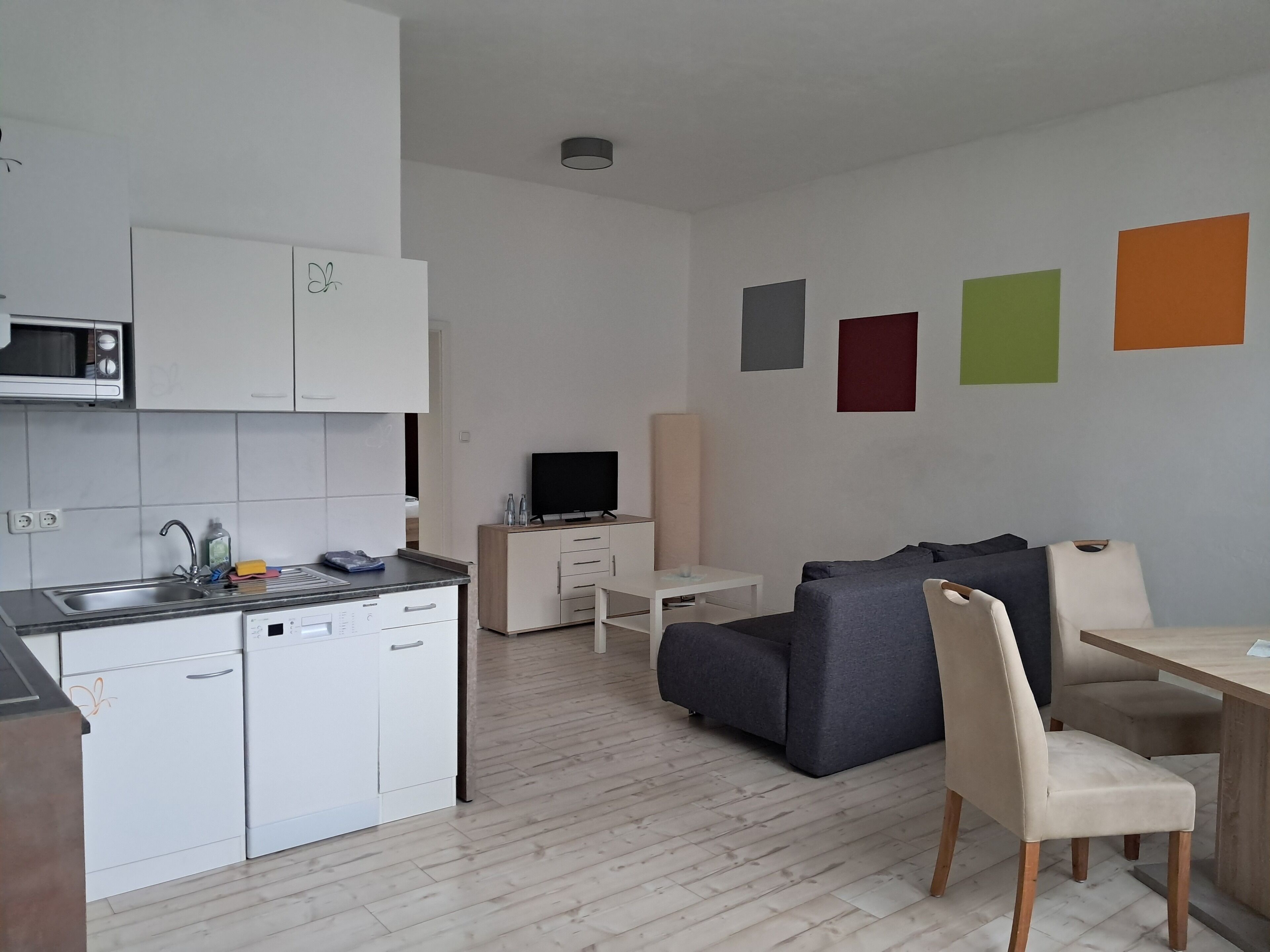 A modern apartment with a kitchen, a grey sofa, a wooden dining table, and a TV.