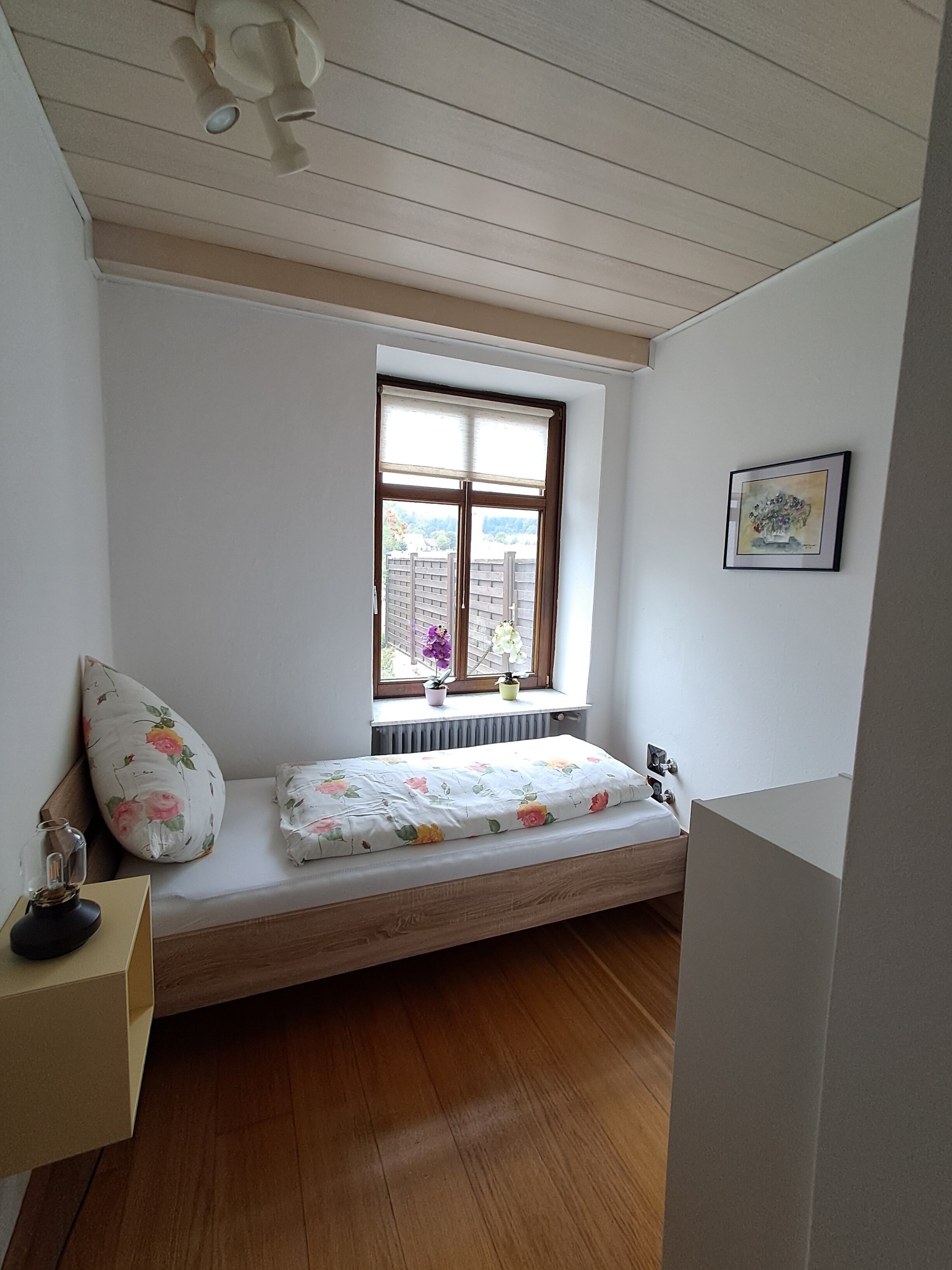 Una habitación pequeña y acogedora con piso de madera, una ventana con vistas, una cama con ropa de cama floral y un cuadro en la pared.