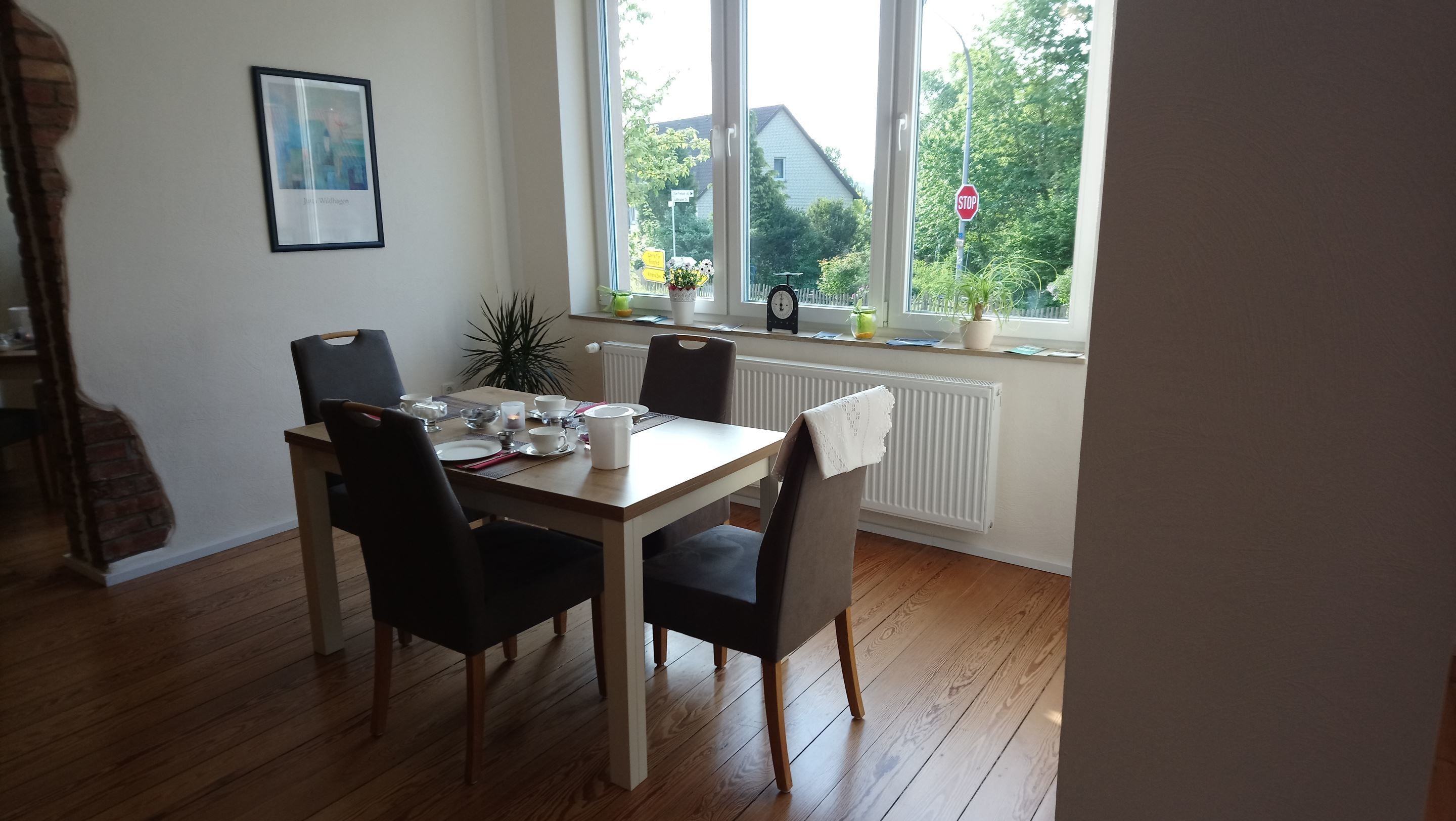 Een eetgedeelte met een tafel gedekt voor vier personen, stoelen, een lijst aan de muur en een raam met uitzicht op huizen en groen.