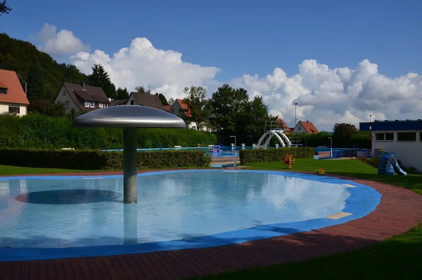 A swimming pool with a central fountain, surrounded by a brick walkway and a playground area.