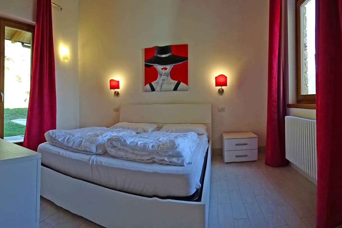 A bedroom with a bed, bedside tables, a painting, and red curtains.