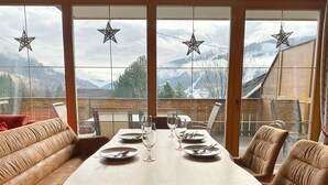 A dining area with a wooden table set for four, leather chairs, and a view of snow-covered mountains through large windows. - Haus Sonnwiese Ski-to-Door (Bad Kleinkirchheim)