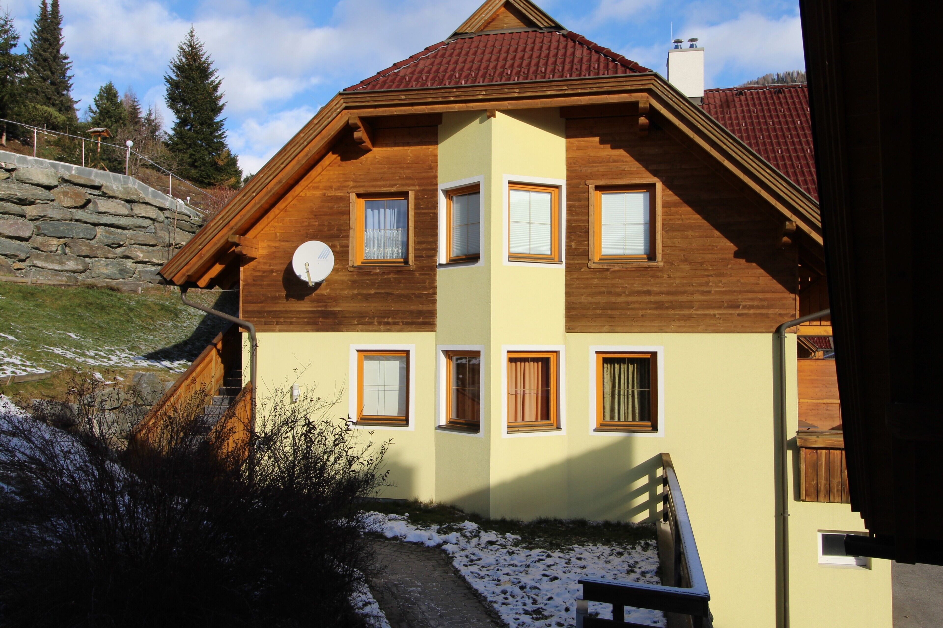 A two-story wooden house with a sloped roof, a stone retaining wall, and a satellite dish.