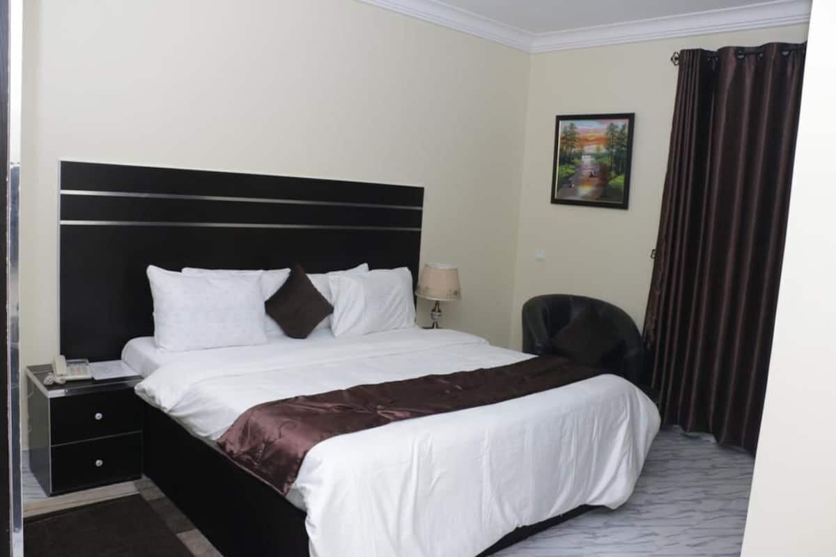 Quarto de hotel com cama, mesa de cabeceira, luminária, cadeira e um quadro na parede.