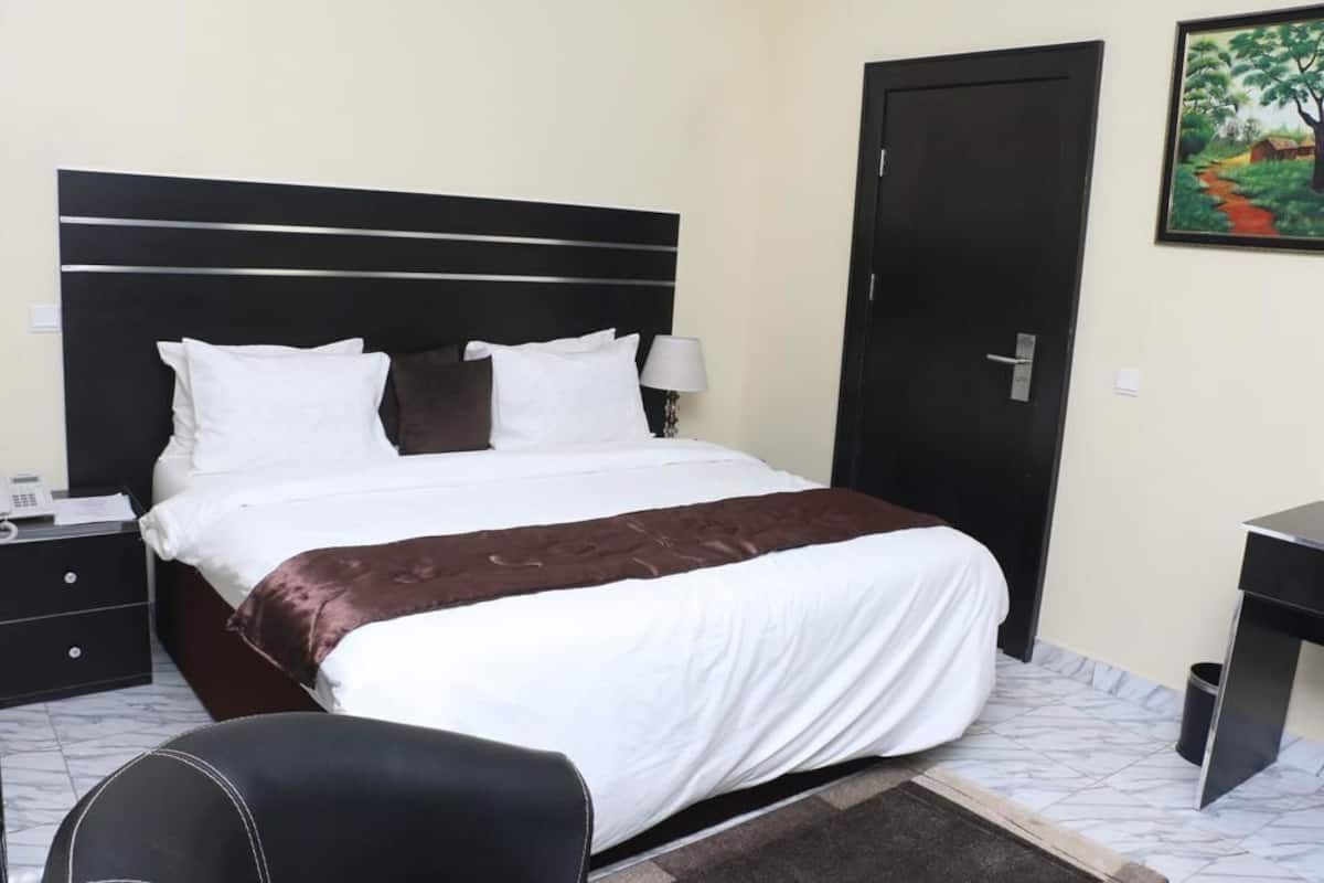 Quarto de hotel com cama, escrivaninha, cadeira, mesa de cabeceira e porta.