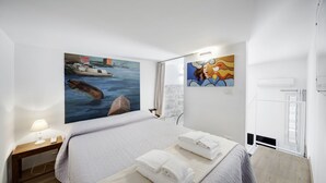 A hotel room with a bed, a nightstand with a lamp, a painting of a boat on the wall, and a window with curtains. - Just 50 meters from one of the most beautiful and renowned beaches of the Ragusa’s coas (Marina di Ragusa)