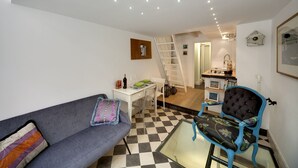 A living room with a grey sofa, a blue armchair, a glass coffee table, a white desk, and a staircase leading to an upper level. - Just 50 meters from one of the most beautiful and renowned beaches of the Ragusa’s coas (Marina di Ragusa)