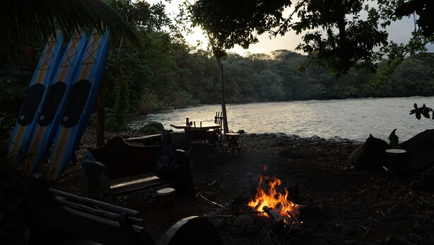 Een camping met een vuur, banken en een tafel onder een boom aan een meer.