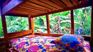 Een bed met een kleurrijk bloemenbedekking, een houten hoofdbord en grote ramen met uitzicht op weelderige begroeiing.