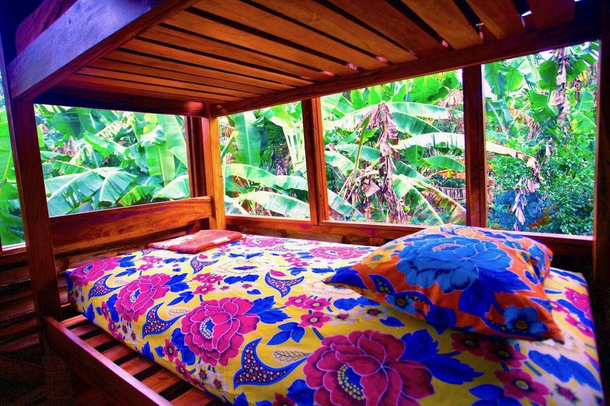Ein Bett mit farbenfroher Blumenbettwäsche, einem Holzkopfende und großen Fenstern mit Blick auf üppiges Grün.