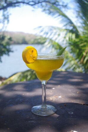 Un verre à cocktail, orné d’une tranche de citron, est posé sur une table. En arrière-plan, on aperçoit un plan d’eau et de la végétation.
