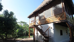Een rustiek houten huis met rieten dak, een houten trap naar een balkon en een pad van stenen.