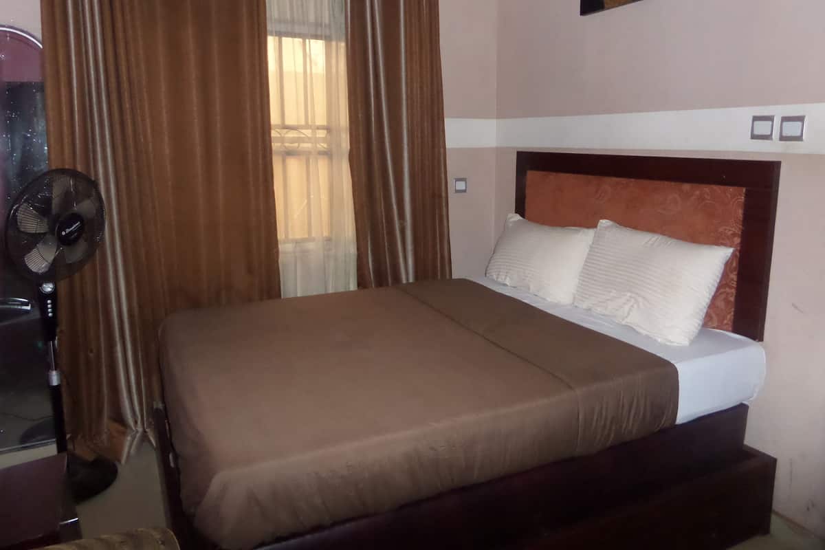 A bedroom with a bed, curtains, a standing fan, and a wall-mounted picture.