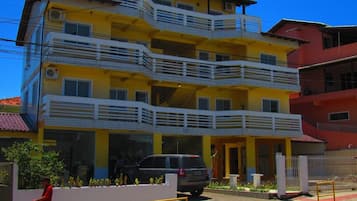 Un edificio amarillo de varios pisos con balcones, un coche estacionado adelante y un peatón caminando.