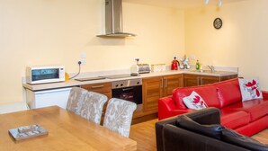 A compact kitchen with modern appliances, a dining area with a wooden table and patterned chairs, and a red sofa with floral cushions. - The Coachhouse (Colwyn Bay)