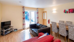 A living room with a TV, a red sofa, a black leather couch, a dining table with patterned chairs, and a door leading outside. - The Coachhouse (Colwyn Bay)