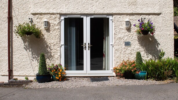 A building with a textured facade, a set of double doors, and potted plants with flowers. - The Coachhouse (Colwyn Bay)