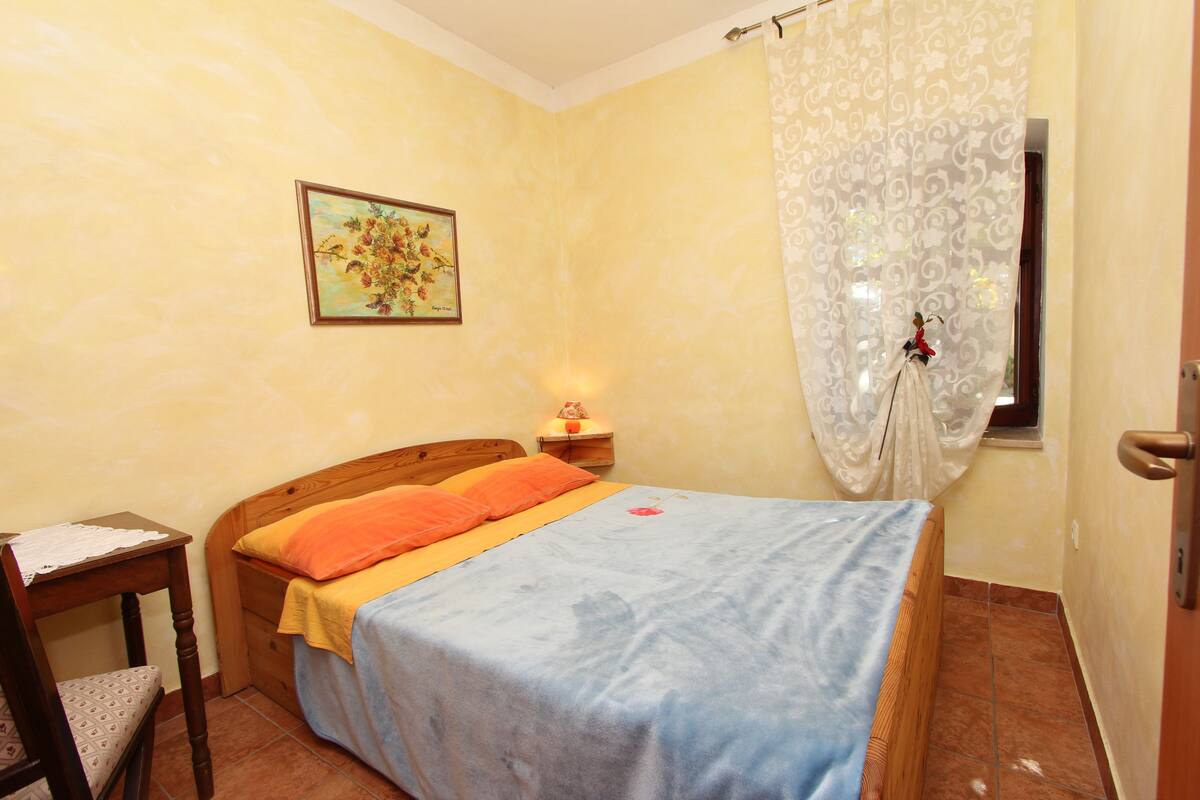 Ein Schlafzimmer mit einem hölzernen Bett, einem Nachttisch mit Lampe, einem Sessel und einem Fenster mit Vorhängen.