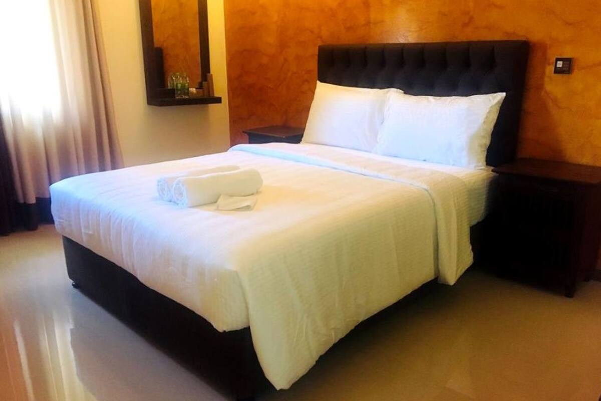 Een net opgemaakt bed met witte lakens en een opgerolde handdoek, een donker hoofdbord en een klein nachtkastje met een lamp.