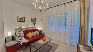 A modern living room with a red sofa, a coffee table, a chandelier, and a large window with sheer curtains. - Apartments Godena (Rovinj)