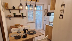 A modern kitchen with a wooden table, white chairs, and a coffee maker. There are hanging light fixtures and a window with a view of trees. - Apartments Godena (Rovinj)