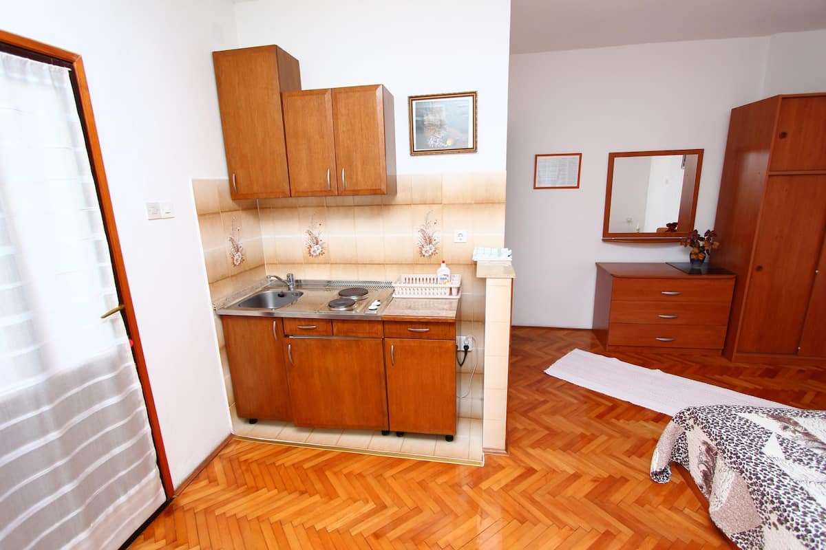 A compact kitchen with wooden cabinets, a sink, and a countertop. There is a mirror above the sink and a wooden wardrobe in the adjacent room.