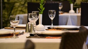 Una mesa de comedor dispuesta con platos blancos, copas de vino transparentes y cubiertos, con un pequeño jarrón blanco y una planta verde al fondo.