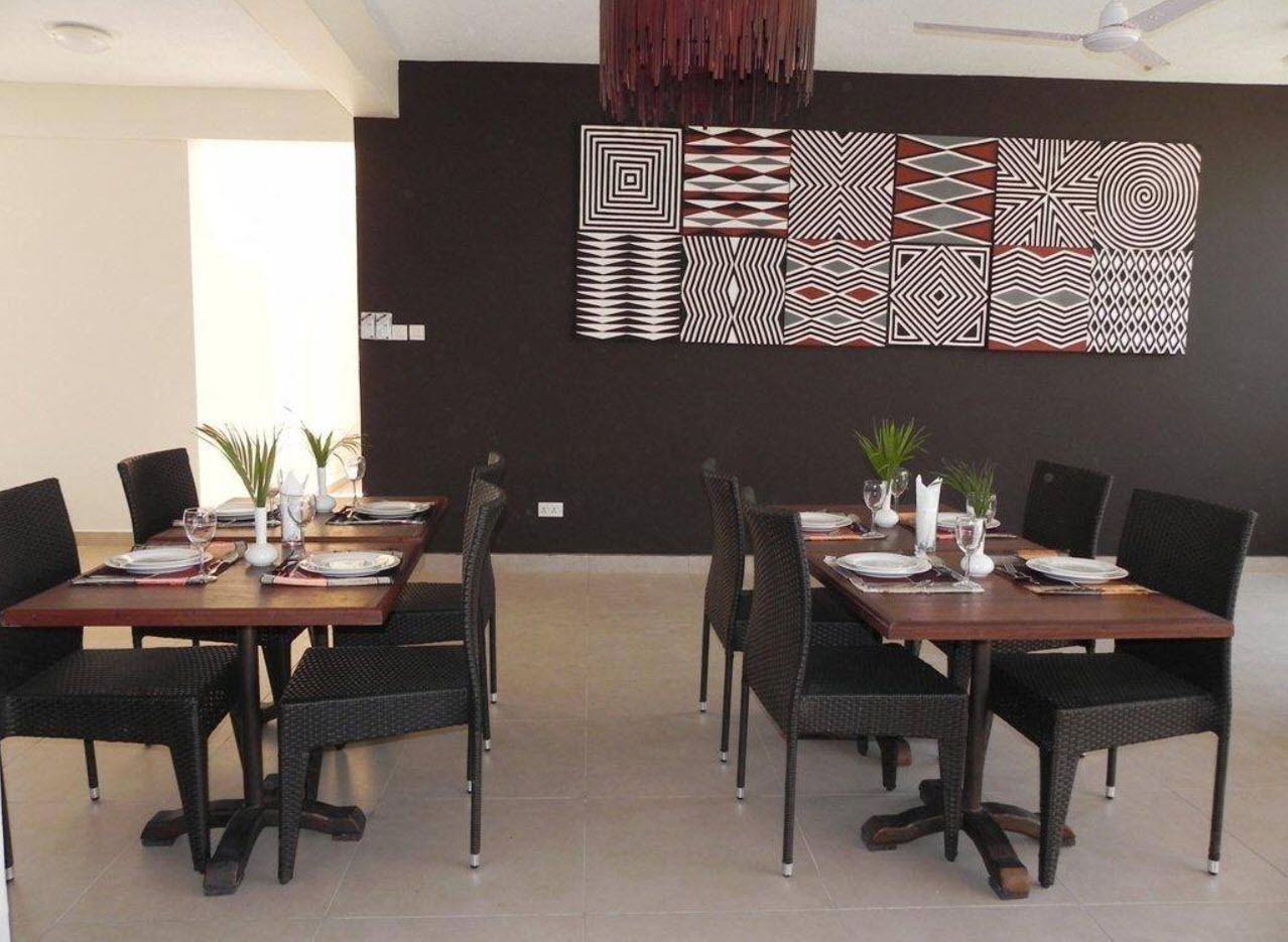 A modern dining area with six tables, each set with plates, glasses, and cutlery. The room features a dark wall with geometric wall art and a ceiling with a hanging light fixture.