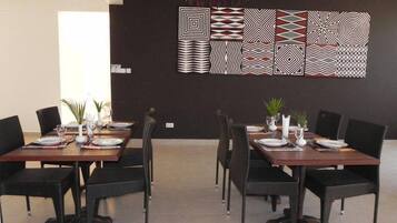 A modern dining area with six tables, each set with plates, glasses, and cutlery. The room features a dark wall with geometric wall art and a ceiling with a hanging light fixture.
