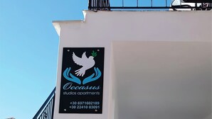 A white building with a sign for 'Occasus Studios Apartments' and contact numbers. - Occasus Room (Halki)