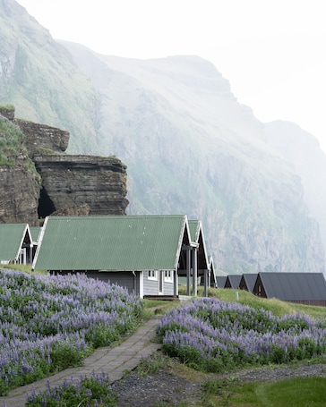 A row of wooden cabins with green roofs, set against a backdrop of mountains and a field of purple flowers.