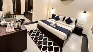 A modern hotel room with a bed, bedside tables, a desk with a chair, a water bottle, a kettle, and a tray with a cup and a thermos. - Hollyhocks Residency (Gurugram)