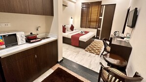 A compact hotel room with a kitchenette, a bed with red bedding, a seating area with a chair, and a small table. - Hollyhocks Residency (Gurugram)