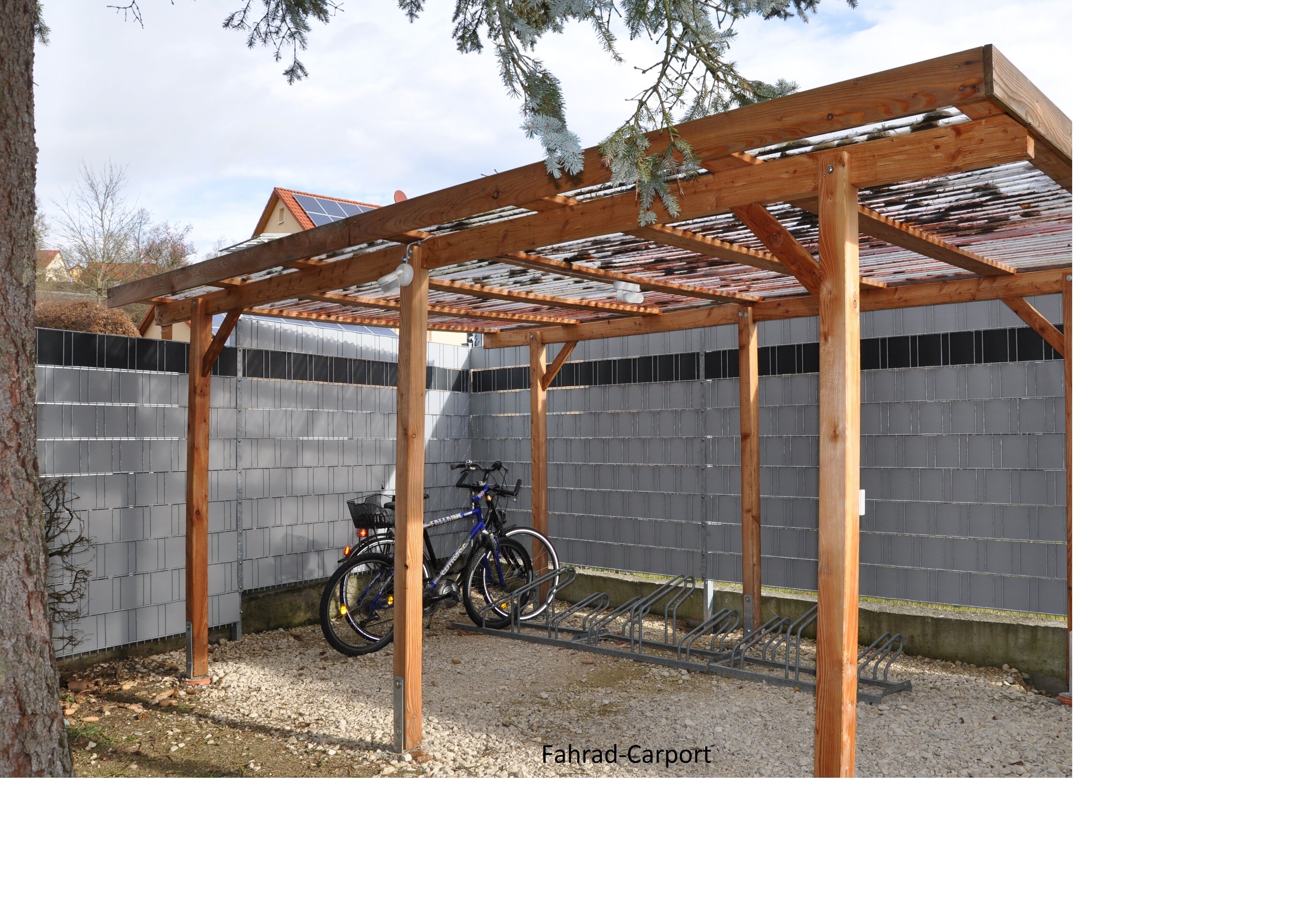 A wooden carport with a bicycle rack, gravel ground, and a metal fence in the background.