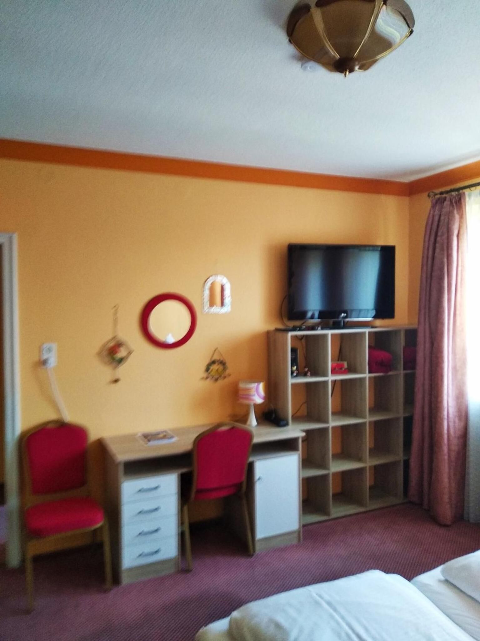 Et hotelværelse med seng, skrivebord med stol, fjernsyn på hylde og et klædeskab.