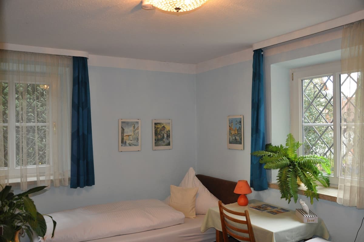A room with a bed, a table with a chair, a potted plant, and two framed pictures on the wall.