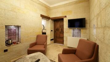 Una habitación de paredes de piedra con dos sillones marrones, una mesa de centro, un televisor montado en la pared y una puerta de madera.