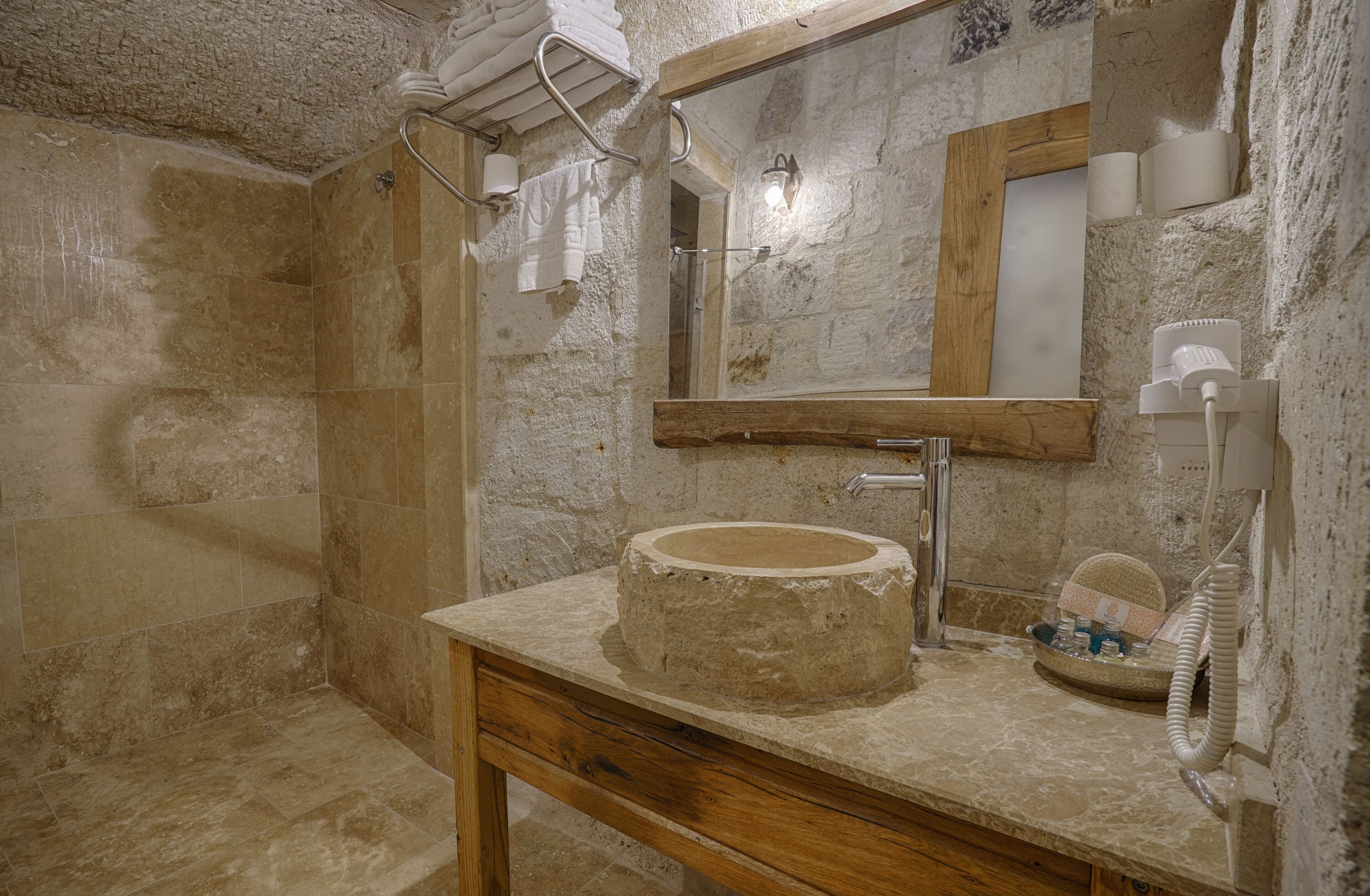 A bathroom with a stone wall, a wooden vanity, a round sink, a mirror, and a shower area.