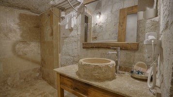 A bathroom with a stone wall, a wooden vanity, a round sink, a mirror, and a shower area.