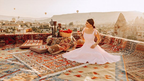 Una mujer con vestido blanco sentada sobre una alfombra estampada, con un paisaje urbano y globos aerostáticos al fondo.