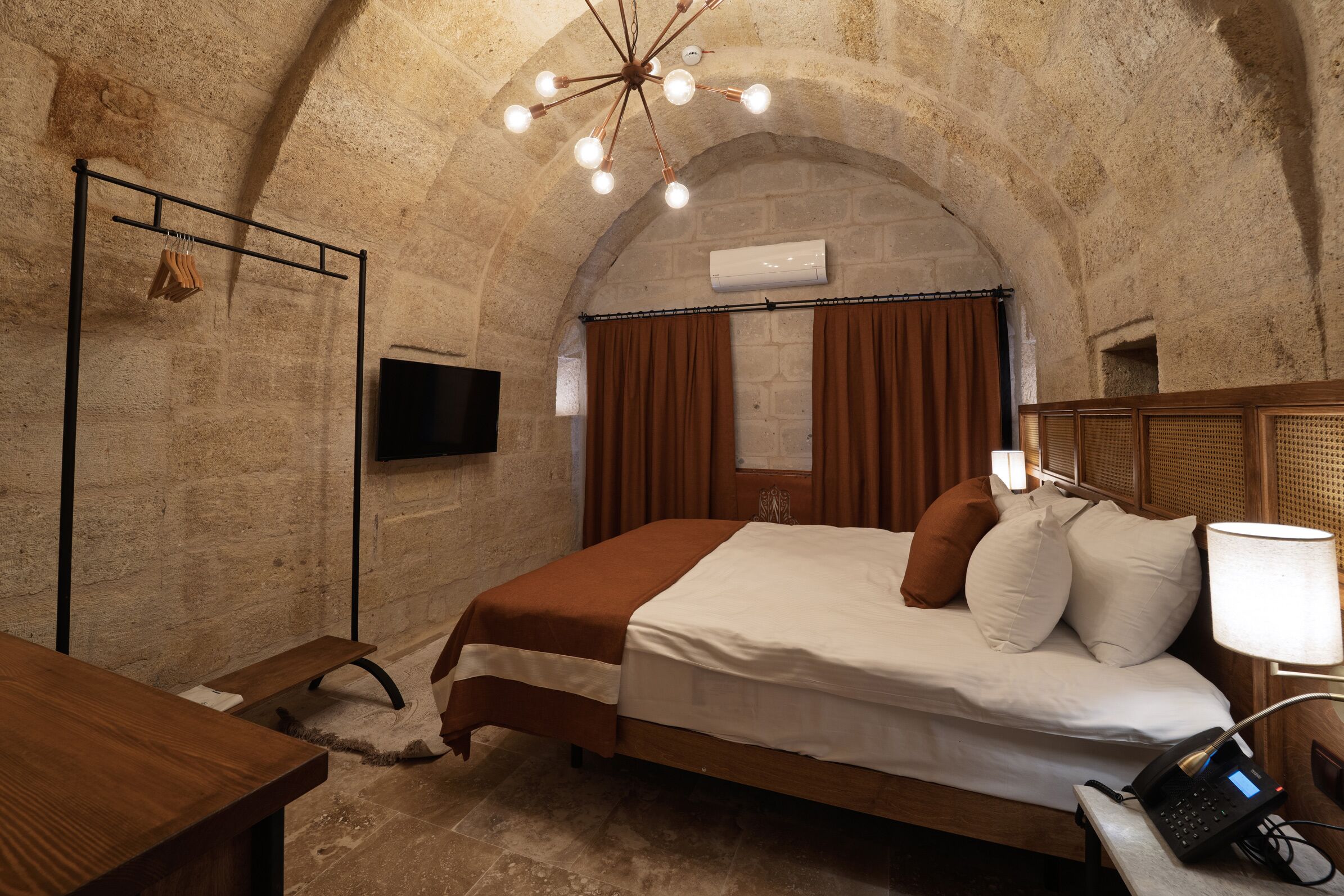 A room with a stone wall, a bed with a brown and white bedspread, a wooden nightstand, a lamp, and an air conditioning unit.