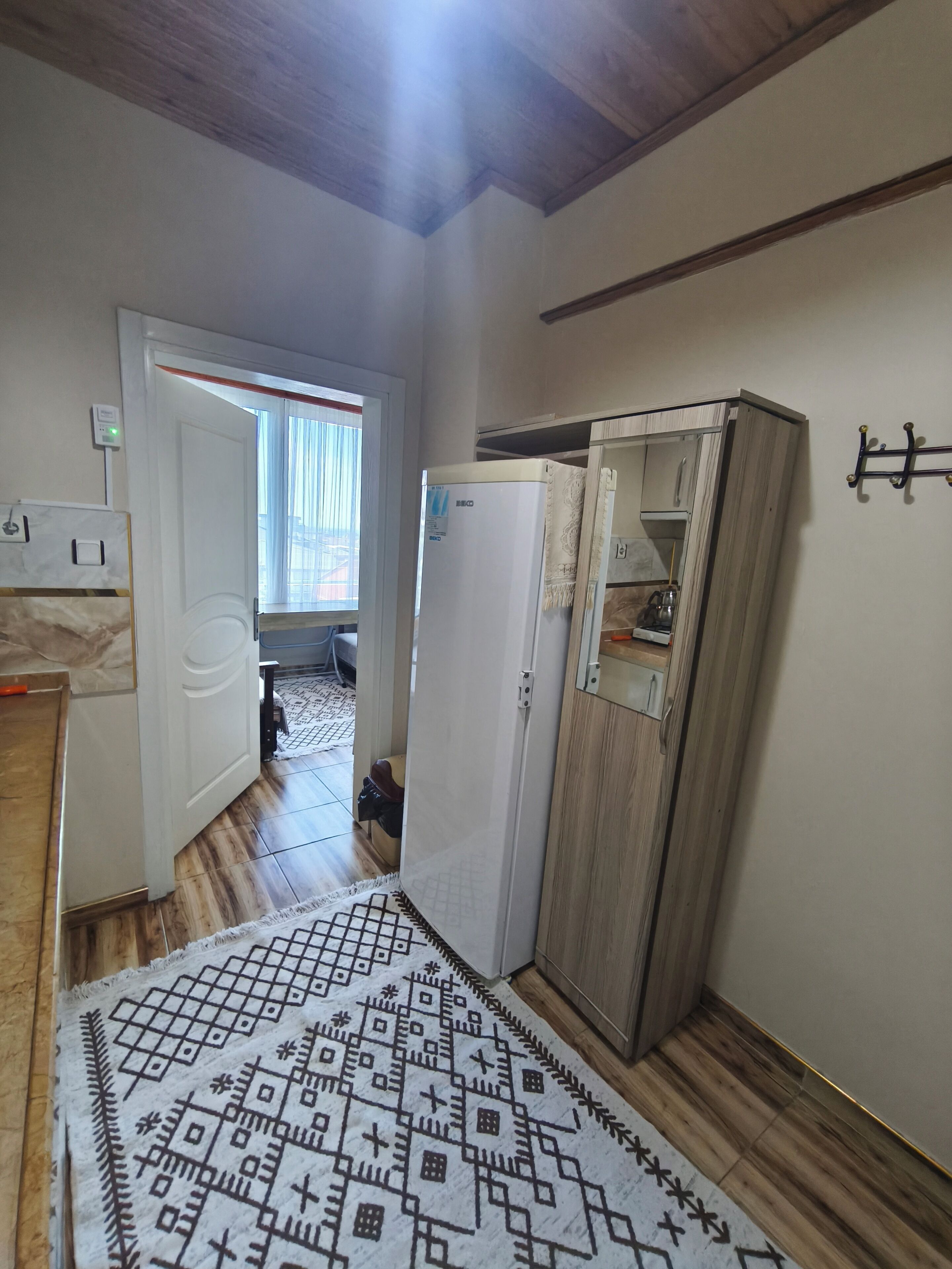 Un pasillo estrecho con un tapete estampado, un armario blanco, un mueble de madera y una ventana con persianas.