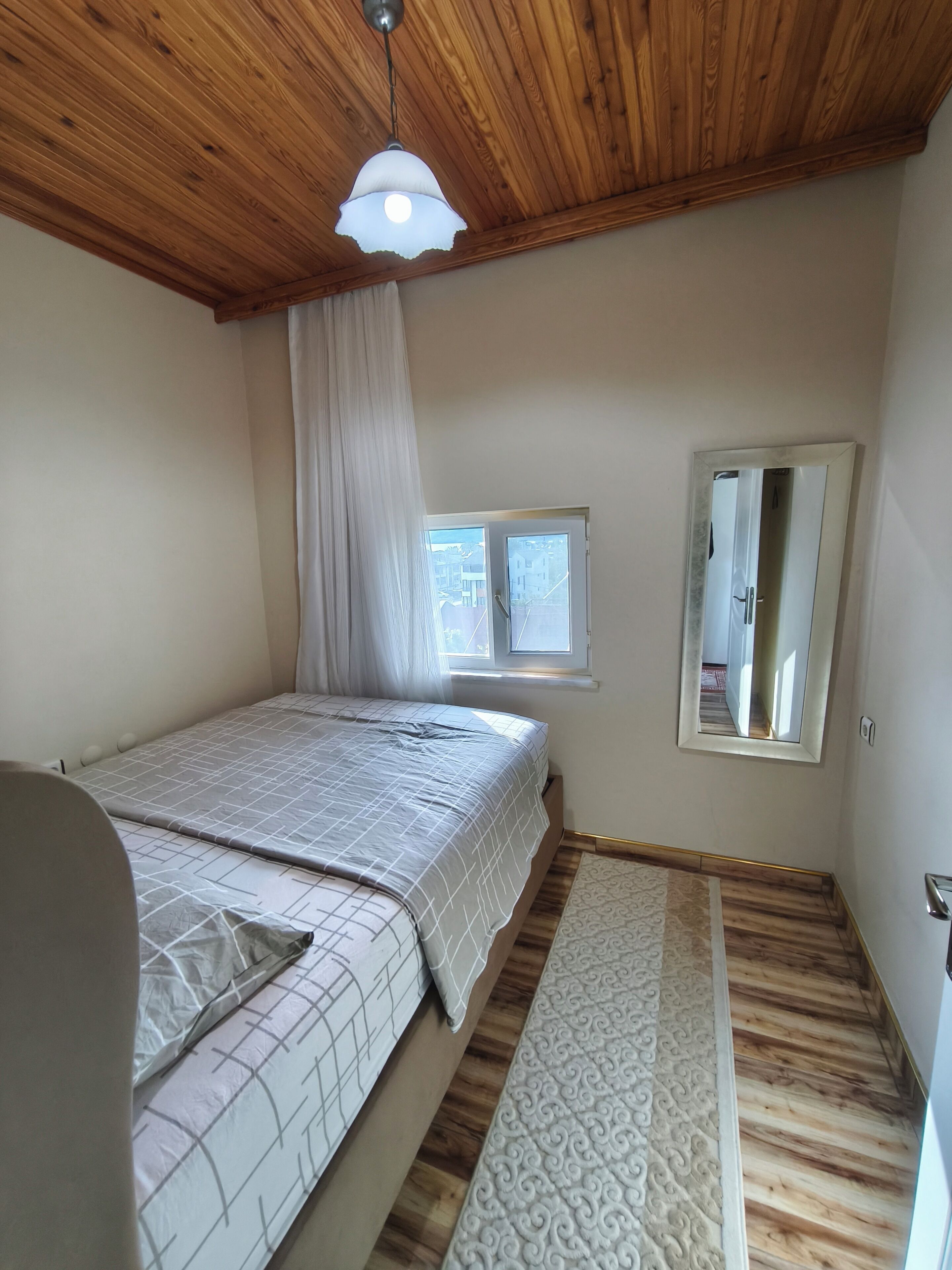 A bedroom with a bed, a window, a mirror, and a wooden ceiling.