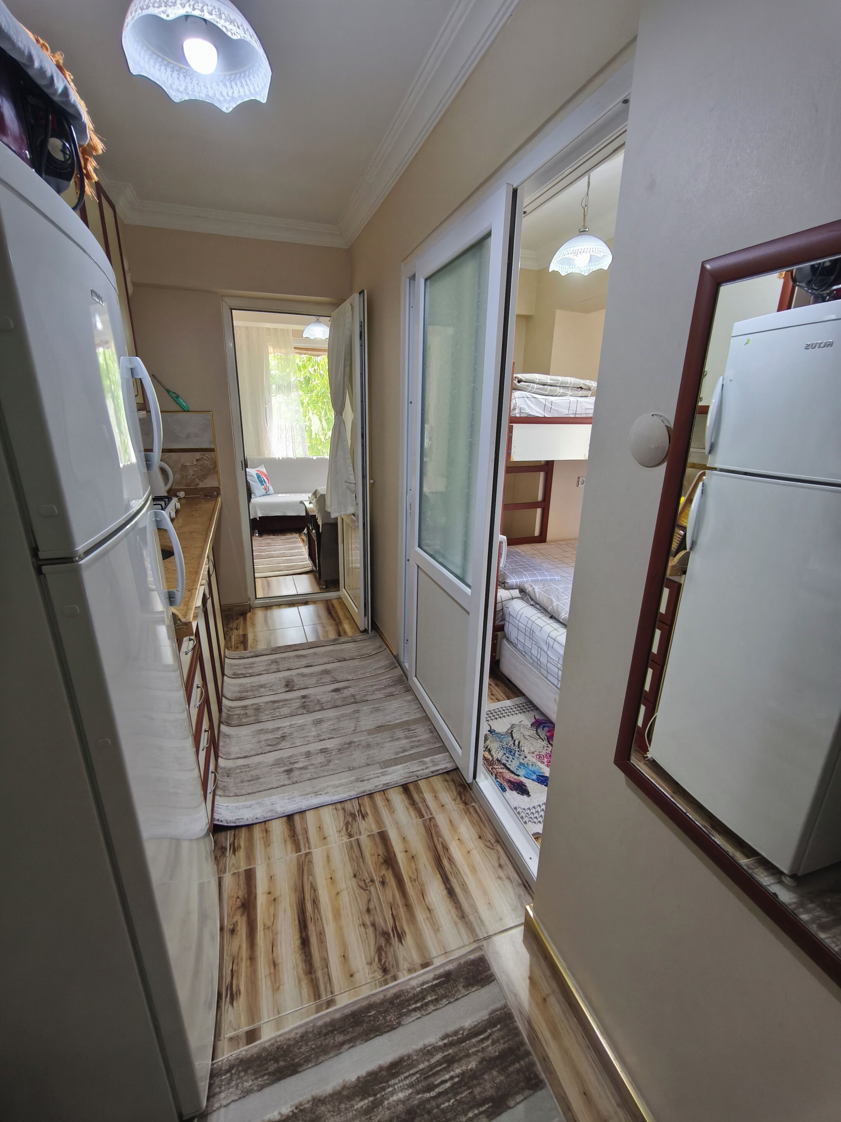 Un pasillo estrecho con suelo de madera, un refrigerador blanco, una puerta corrediza que da a una habitación con cama y una lámpara en el techo.