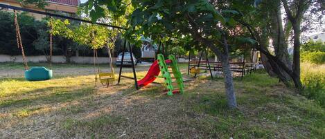A playground with swings and a slide, surrounded by trees and grass.
