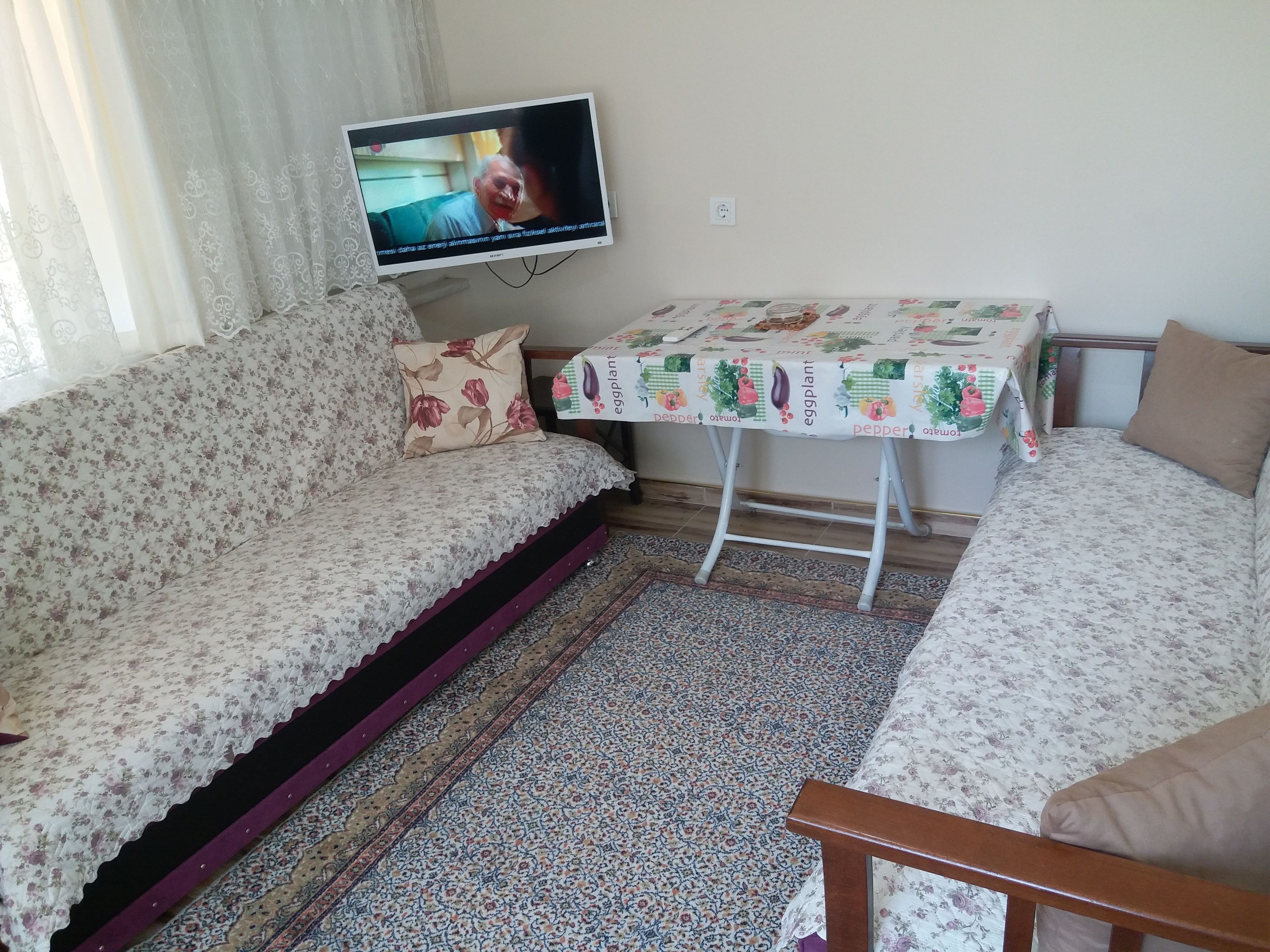 A living room with a sofa, a small table with a tablecloth, a television, and a patterned rug.