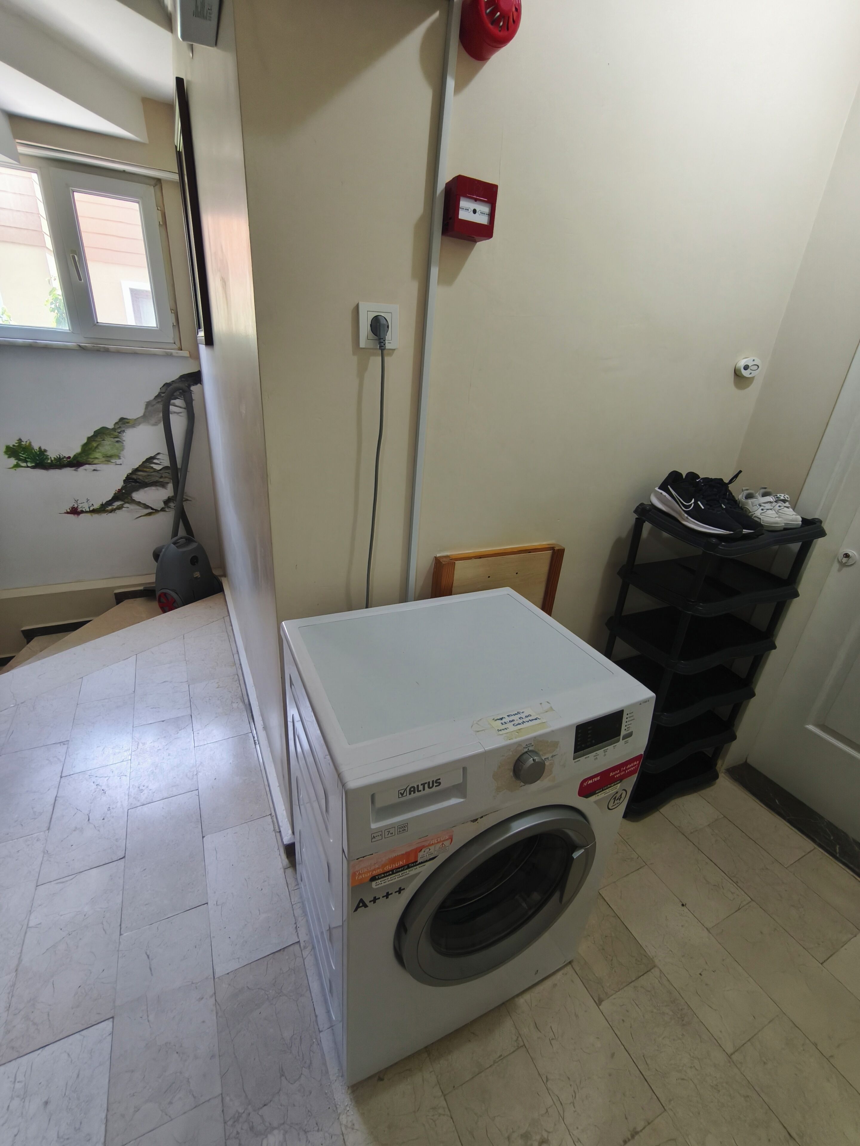 A compact laundry room with a front-loading washing machine, a small table, a shoe rack, and a window.