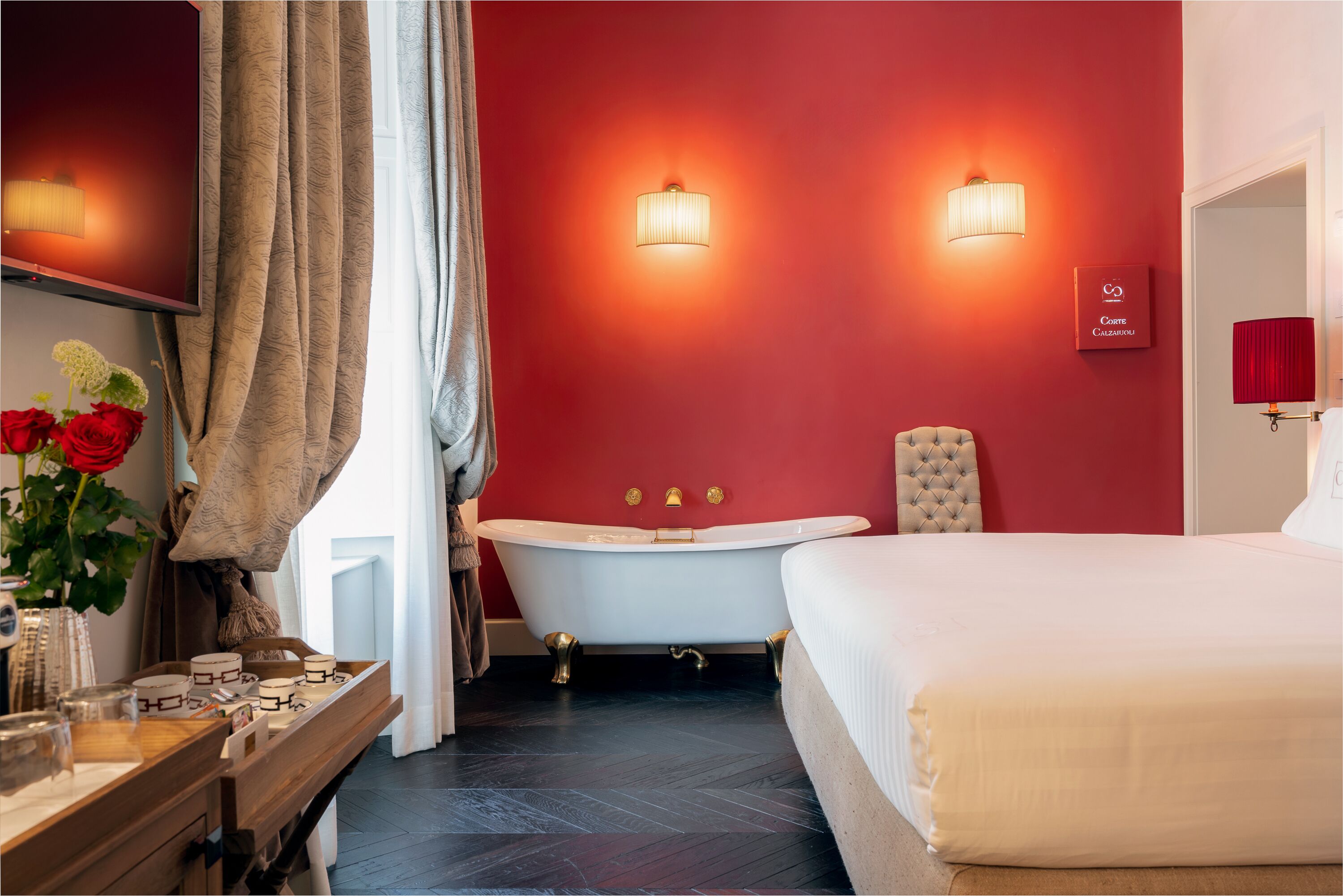 A modern bathroom with a freestanding bathtub, a vanity with a mirror, and a bedside table with a vase of red roses.
